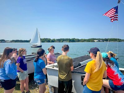 High schoolers crowd around large vats near the shoreline with a sailboat on the water behind them